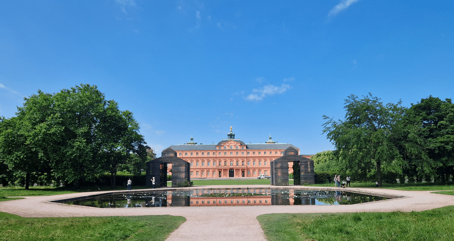 Château de Rastatt avec bassin Château de Rastatt, bâtiment baroque rouge avec dôme central, se reflète dans un bassin rectangulaire. Pelouses vertes et arbres matures encadrent la scène sous ciel bleu.