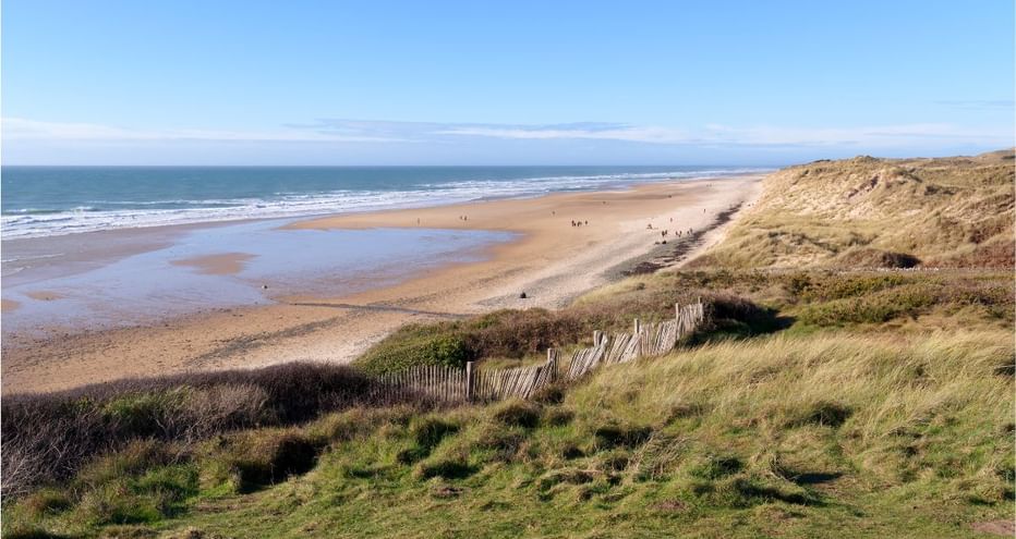 Large plage de sable à Hatainville avec dunes herbeuses, clôtures en bois et mer bleue calme sous ciel dégagé dans le Cotentin, France.
