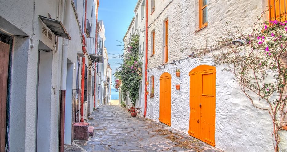 Ruelle pavée étroite à Cadaqués avec bâtiments blanchis à la chaux et portes et fenêtres orange vif, menant vers la mer sous ciel bleu.
