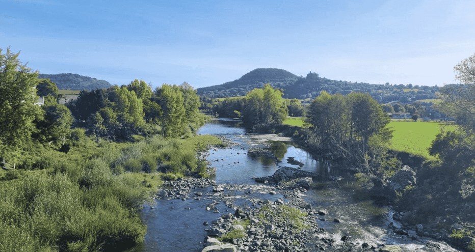 Rivière rocailleuse traversant un paysage verdoyant avec des arbres sur les deux rives. Collines et montagnes lointaines sous ciel bleu.