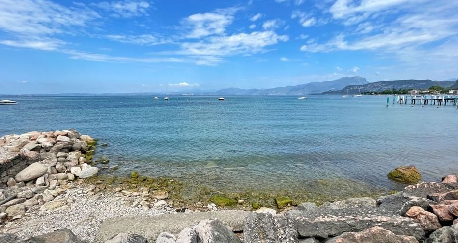 Rivage rocheux du lac de Garde près de Bardolino avec eau bleue claire, brise-lames en pierre et montagnes en arrière-plan sous ciel nuageux.
