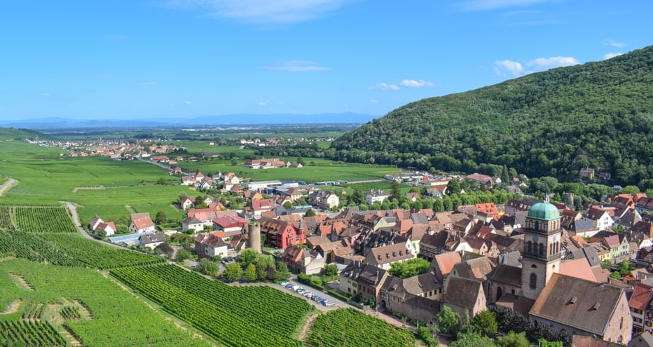 Vue aérienne du village de Kaysersberg en Alsace avec maisons traditionnelles, clocher, entouré de vignobles verts et collines boisées.