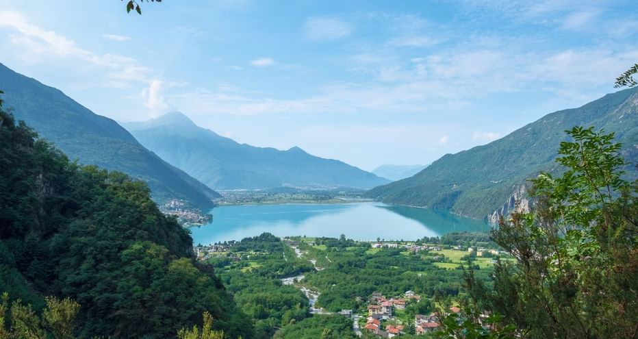 Lac de Mezzola niché dans une vallée entre des montagnes vertes. Un village avec des maisons au premier plan, entouré de végétation luxuriante.