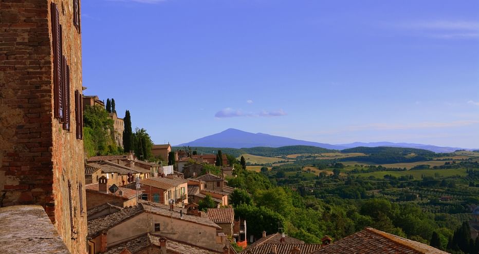 Vue de Montepulciano montrant des toits en terre cuite, des cyprès et des collines toscanes avec une montagne lointaine sous un ciel bleu.