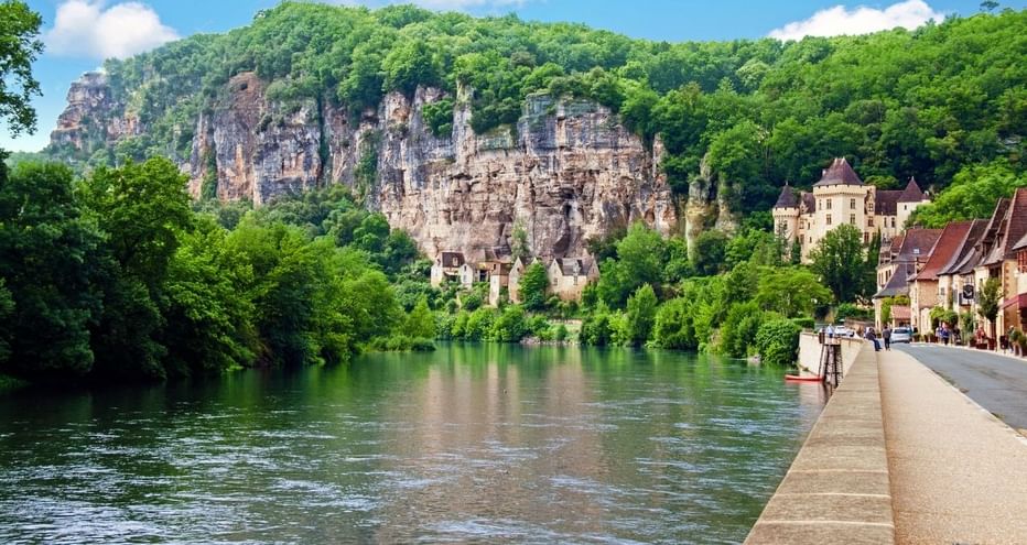 Rivière longeant des maisons en pierre en Périgord avec falaises calcaires et forêt verte. Une promenade borde l'eau sous un ciel bleu.