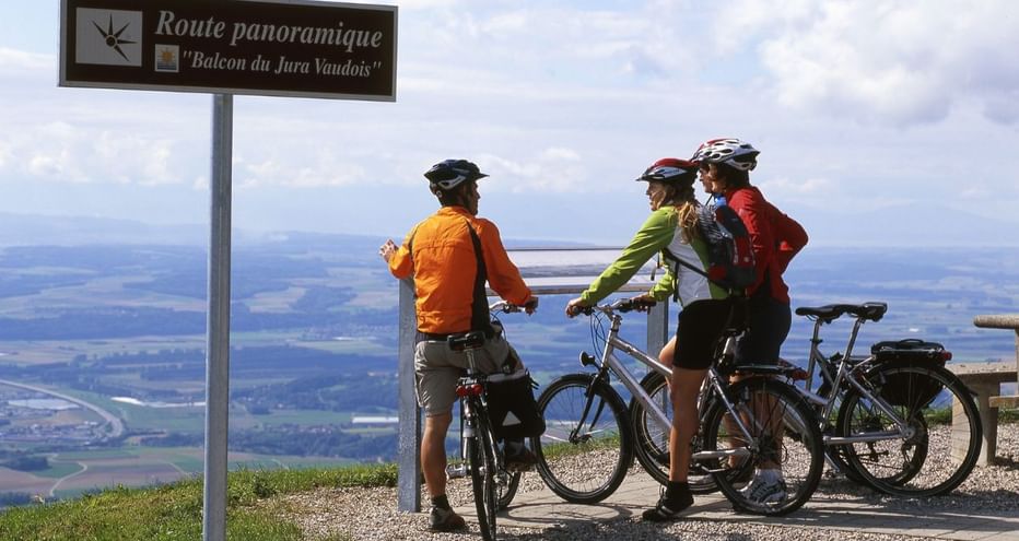 Trois cyclistes avec casques et sacs à dos au point de vue panoramique Balcon du Jura Vaudois, surplombant le paysage de vallée suisse.