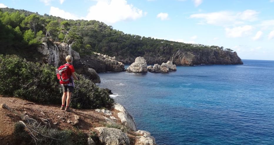 Randonneur avec sac à dos rouge sur falaise rocheuse de Majorque, contemplant les eaux turquoise de la Méditerranée et falaises calcaires.