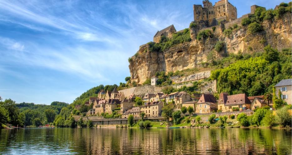 Village médiéval avec château en pierre perché sur falaise calcaire au-dessus de la Dordogne calme en Périgord. Maisons traditionnelles en cascade vers l'eau.