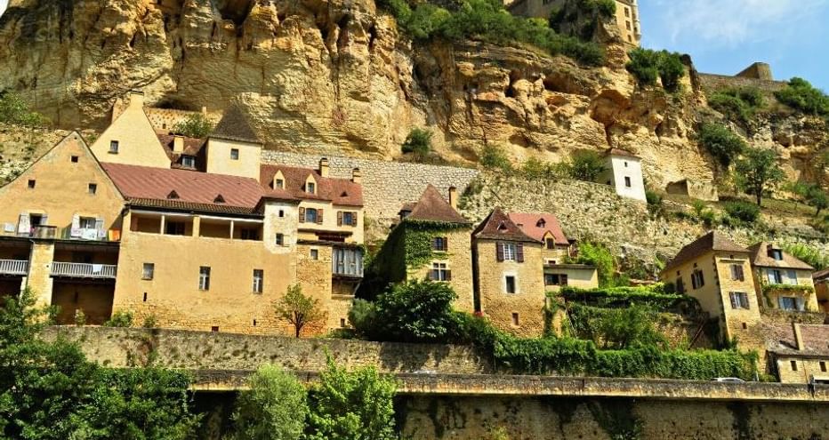 Maisons en pierre aux toits de tuiles construites le long d'une falaise calcaire spectaculaire en Périgord Noir, avec ruines de château en haut.