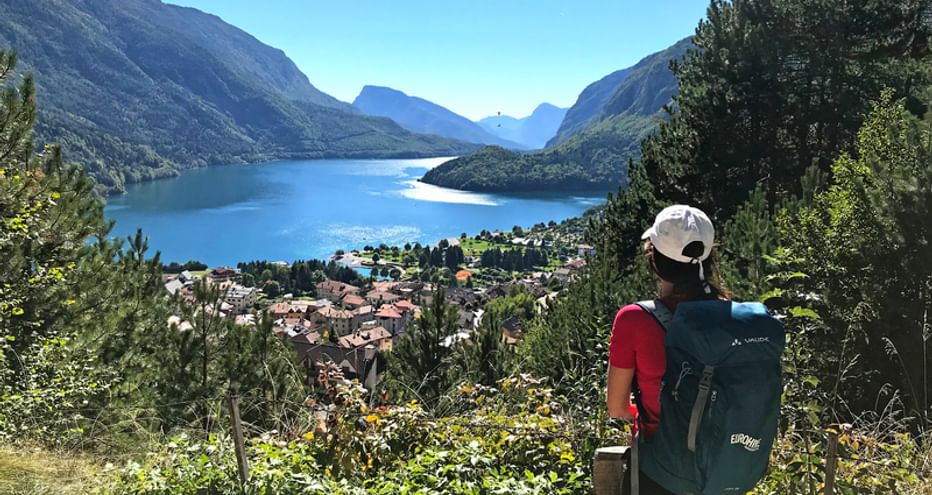 Randonneur avec sac à dos et casquette admirant le lac de Molveno depuis un point de vue élevé, avec montagnes et village au bord du lac bleu.