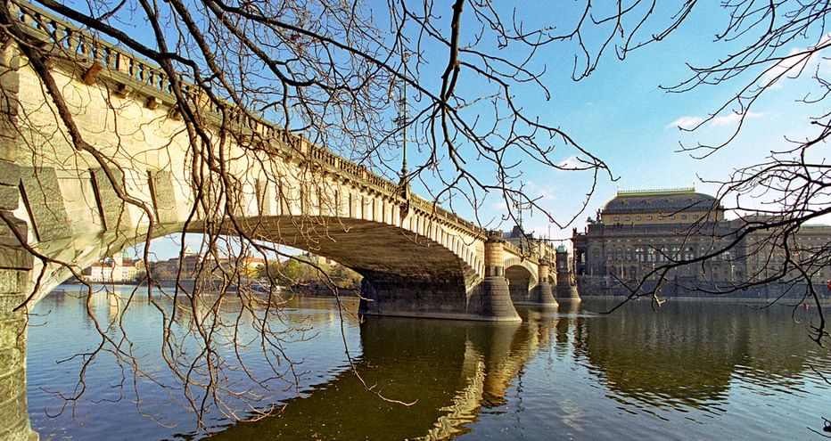 Théâtre national de Prague sur la Vltava Pont en pierre avec arches enjambant la Vltava à Prague, avec le théâtre national visible sur la rive droite sous des arbres nus d'hiver.
