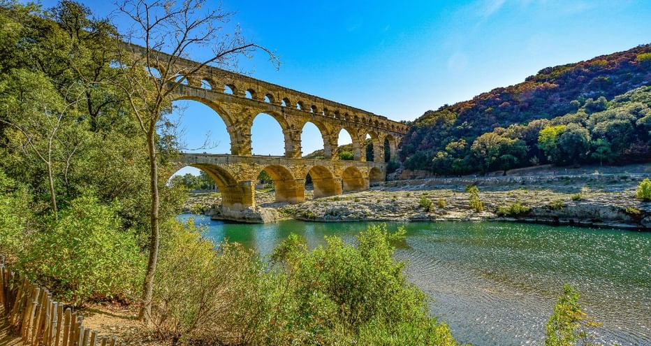 Ancien aqueduc romain du Pont du Gard avec trois niveaux d'arches enjambant le Gardon, entouré de collines vertes et végétation.