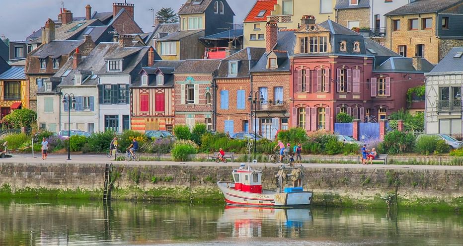Rangée de maisons colorées à Saint-Valéry avec deux petits bateaux amarrés le long d'un quai en pierre. Des gens se promènent sur le front de mer.