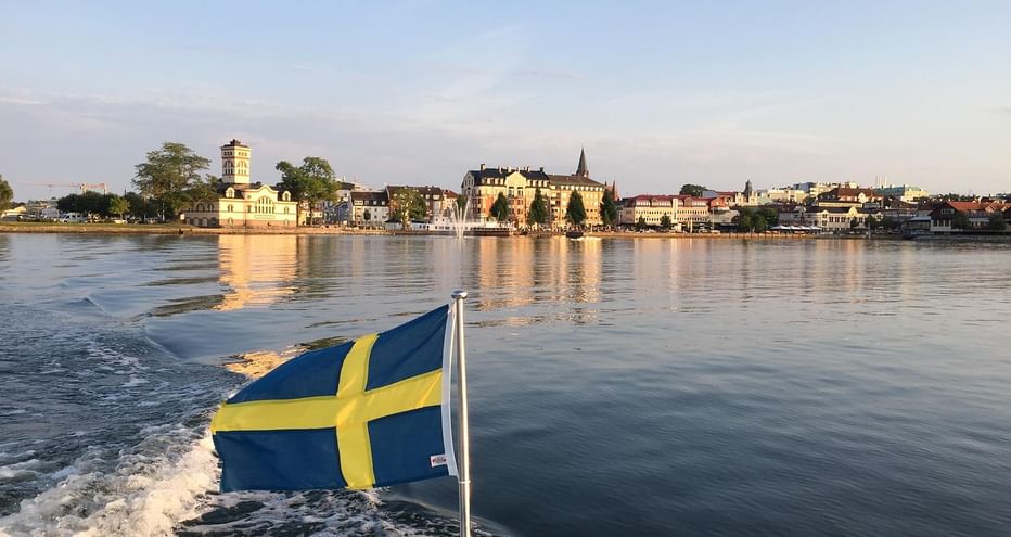 Drapeau suédois flottant sur un bateau avec le front de mer de Stockholm en arrière-plan. Bâtiments historiques et fontaine visibles le long du rivage.