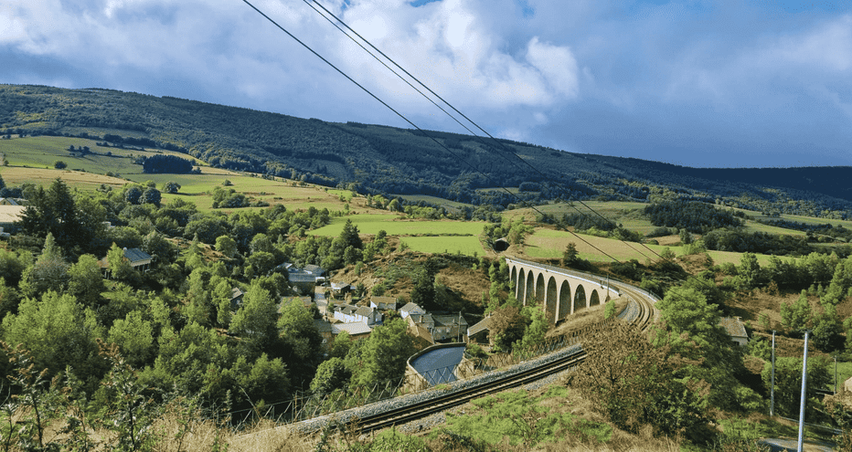 Viaduc ferroviaire en pierre traversant la campagne verdoyante du chemin Stevenson. Collines et forêts sous un ciel bleu avec nuages.