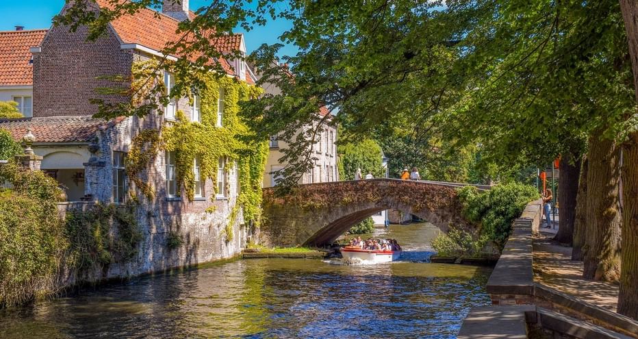 Canal pittoresque à Bruges avec bâtiments médiévaux couverts de lierre, pont de pierre et bateau touristique. Promenade bordée d'arbres.
