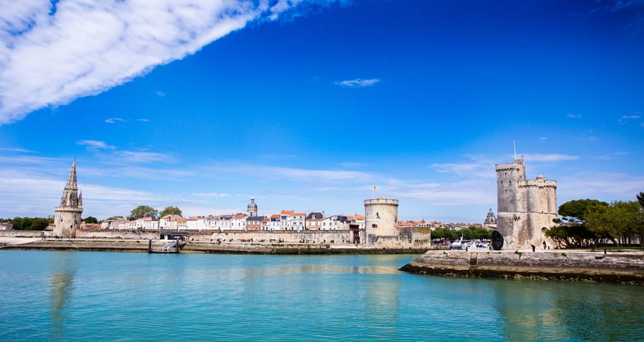 Vue du port de La Rochelle avec eau turquoise, tours médiévales en pierre à droite et bâtiments blancs avec clocher sous ciel bleu.