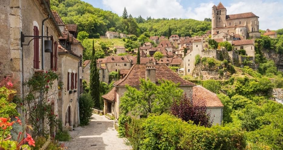 Rue pavée à Saint-Cirq-Lapopie avec maisons médiévales et église sur la colline. Verdure luxuriante autour des bâtiments aux toits de tuiles.