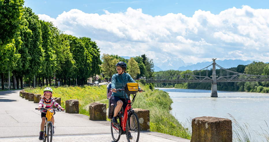 Adulte et enfant à vélo sur chemin riverain à Salzbourg avec la Salzach, pont et montagnes en arrière-plan sous ciel bleu nuageux.
