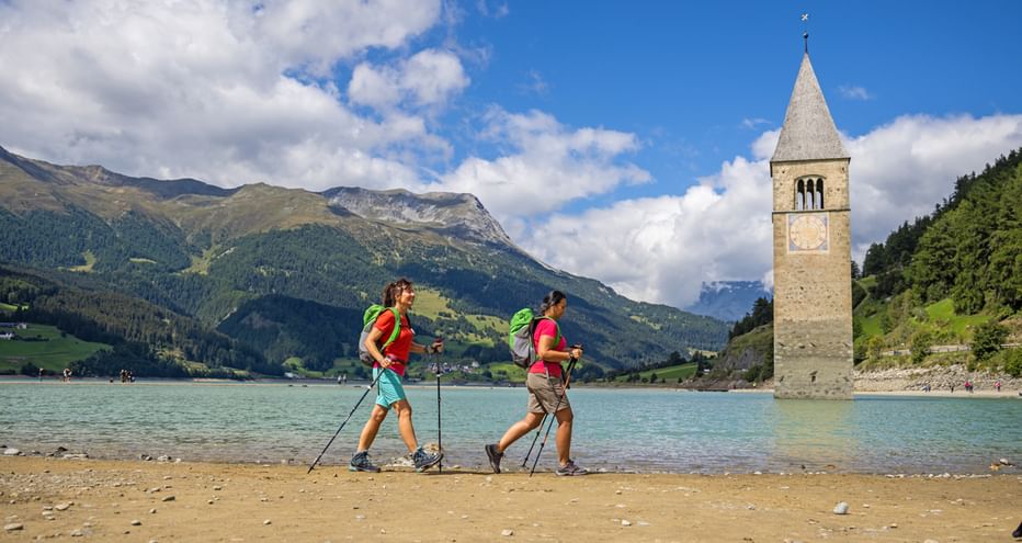 Deux randonneurs marchent sur les rives du lac de Resia, devant le clocher englouti