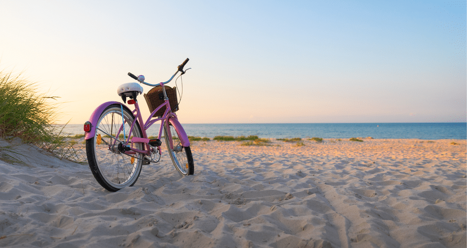 Vélo violet avec panier garé sur plage de sable de la mer Baltique au coucher du soleil, avec herbe de dune et eau bleue calme.
