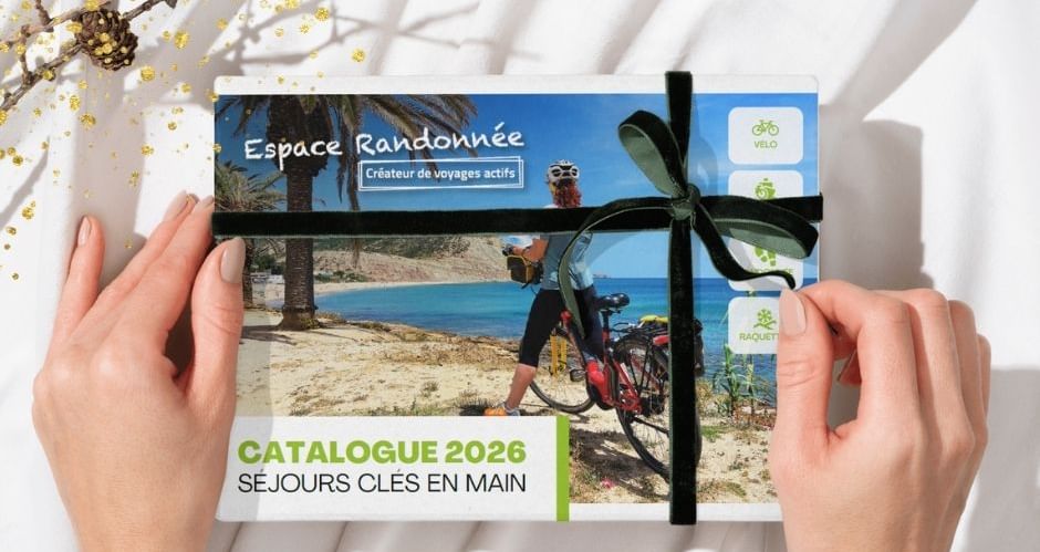 Mains tenant le catalogue Espace Randonnée 2026 avec ruban noir, montrant un cycliste sur la plage. Texte : 'Notre catalogue 2026 bientôt là'.