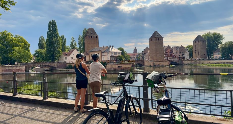 Deux cyclistes avec vélos admirant des tours médiévales de l'autre côté d'une rivière sous un ciel nuageux. Tours en pierre et pont historiques en arrière-plan.