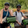 Romain et Vinciane debout avec des vélos équipés de sacoches de guidon vertes, portant casques et lunettes sur un chemin ensoleillé avec des fleurs.